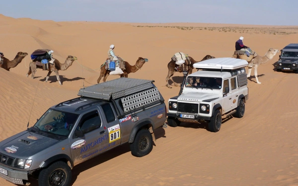 Eine Karawane auf Kamelen kreuzt den Weg der Geländewagen am Rand eines kleinen Dünenbandes.